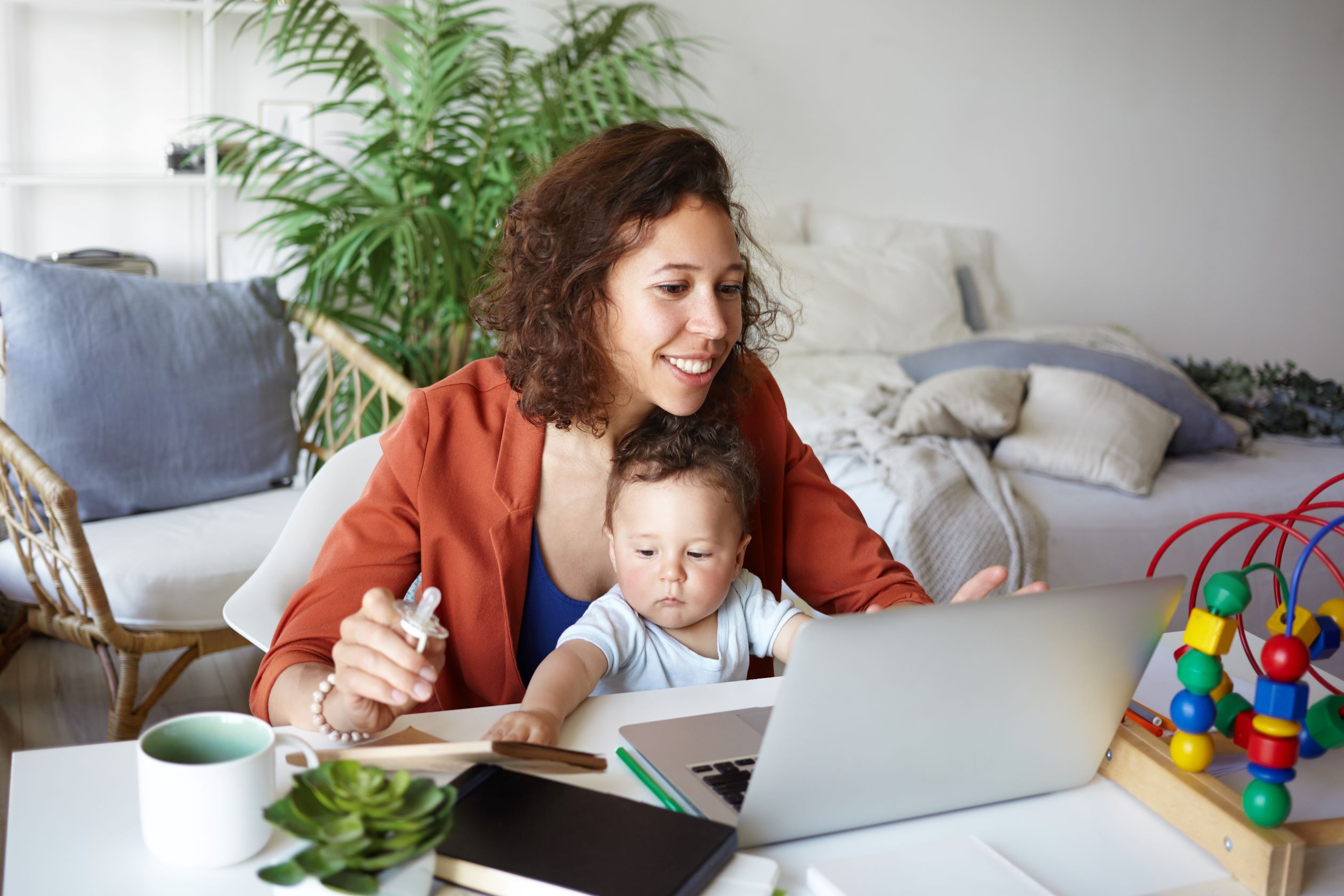 Person working at desk at home using laptop, holding baby on her lap